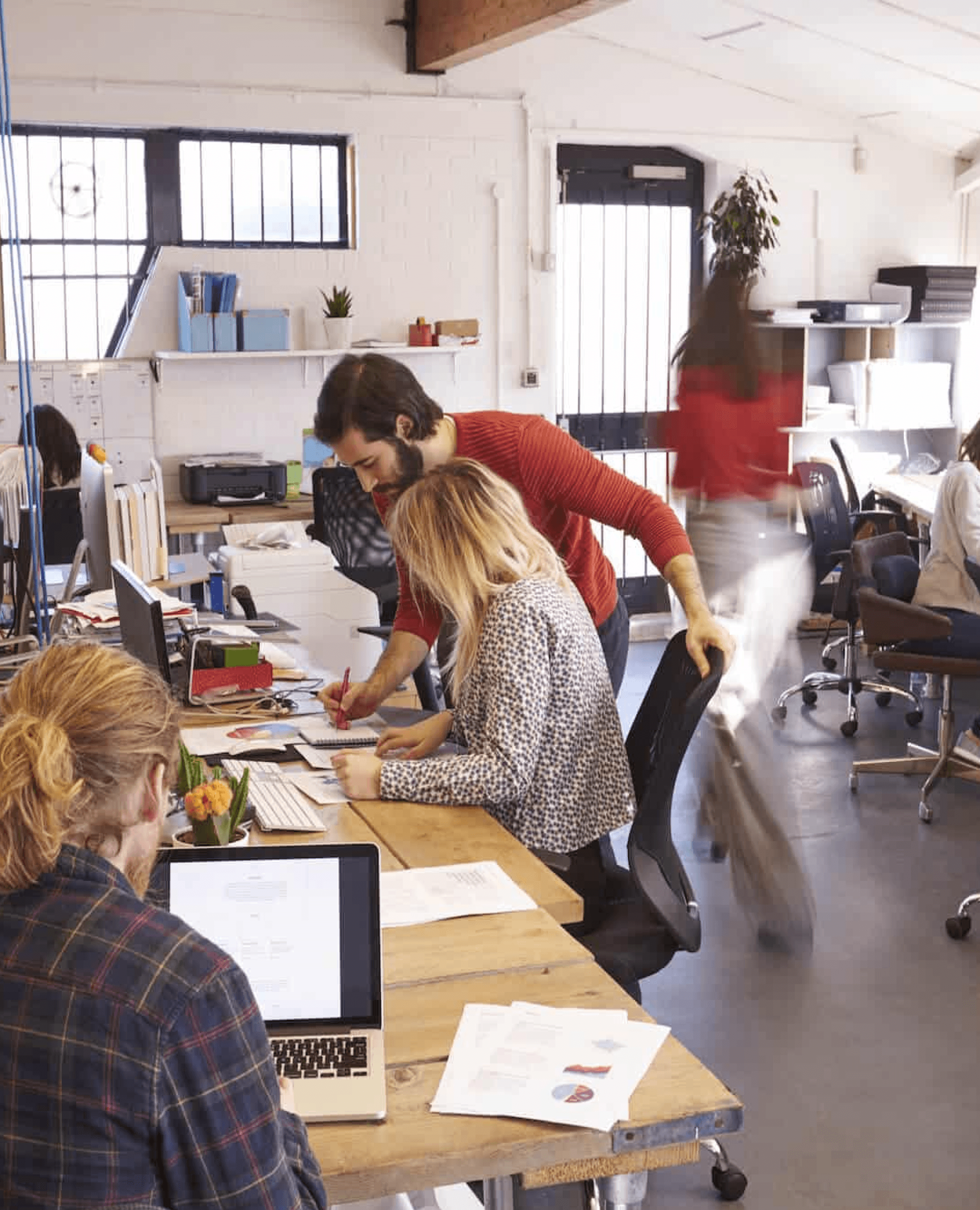 A product team at work in an open studio office: a designer leans over a colleague's desk and they mark up printed design work together.