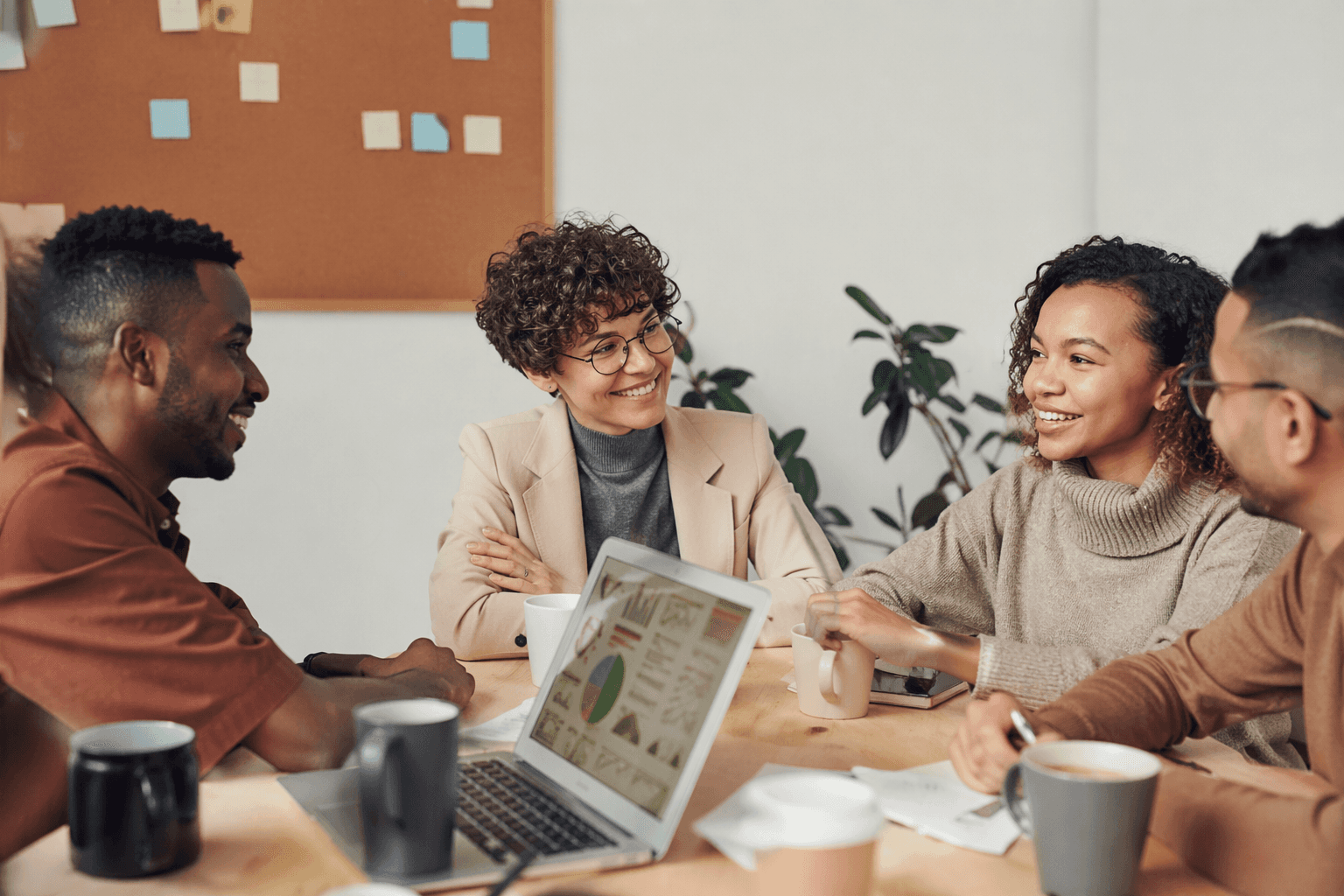 Four colleagues laugh together around a meeting table over coffee, with charts open on a laptop between them.